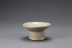 A short glazed and speckled gray porcelain offering dish for an altar. The base is a wide, slightly tapered cylinder which at the top is attached to a shallow dish. At the bottom of the base as well as a circle on the top of the dish there is some discoloration and morphing of the ceramic.<br />
<br />
This dish is gray in color and has large amounts of ash and pinholes on the surface. It is a low-grade object, with sand spur makrs remaining on the inner base due to fired among a stack of other vessels. Coarse sand is stuck to the foot.<br />
[Korean Collection, University of Michigan Museum of Art (2014) p.198]