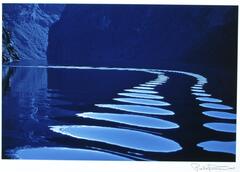 A photograph of ripples on the surface of a calm body of water. A circular pattern curves into the distance, a rocky wall visible in the background. The image is comprised of blue tones.