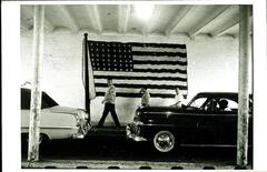 This photograph depicts a cement interior with individuals on foot and in cars passing before a large American flag mural painted on the wall.