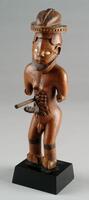 This &nbsp;figure stands in a dynamic pose with knees slightly flexed. This androgynous figure sports a European-style hat, a crescent-shaped beard, and a realistically carved penis. The figure has gracefully curved shoulders and powerful legs. The&nbsp;feet and the arms, originally bent at ninety degrees, have been damaged. Most Bwende figures bear forms of scarification; this figure is no exception. Ornate scarifications are carved in relief upon its abdomen and torso while lined scarifications appear across the chest and forehead and beneath the eyes, which are inlaid with white shell. Most strikingly, however, are the two large metal rods that forcefully pierce the figure&#39;s abdomen.