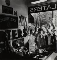 Image of people looking into a bookstore through a large window. The image is taken from the interior and displays University of Michigan memorabilia.
