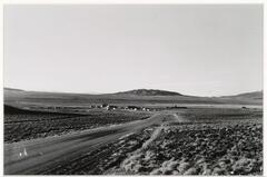 A photograph of a flat landscape bisected by a dirt road, with a settlement in the middle ground and a small mountain visible along the horizon line.