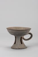 This vessel consists of a cup with an outward-flaring mouth supported on an inverted V-shaped pedestal foot. A single oval-shaped handle is attached to the underside of the cup. The pedestal foot is decorated with four vertical rectangular perforations. Immediately below this is a pair of thin horizontal ridges, which also encircle the body of the cup.<br />
<br />
This is a gray, high-fired stoneware stem cup. It is characterized by its shallow cup body and trumpet-shaped flared pedestal. A raised-band encircles the lower part of the pedestal, which is perforated by rectangular apertures in four places. A loop-shaped handle is attached to the lower part of the cup.
<p>[Korean Collection, University of Michigan Museum of Art (2017) p. 66]</p>
