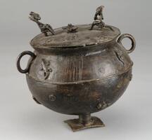 Cast vessel with a round body attached to a rectangular base by a short, spiral column. The body is decorated with figures of turtles, crocodiles, and fish. There are also small spirals across the vessel body. Near the top are two small, round handles. 