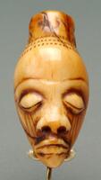 This small Pende ivory pendant (<em>ikhoko</em>) depicts a delicately carved male head. Stylistically, <em>ikhoko</em> bear many of the facial characteristics of anthropomorphic Pende <em>mbuya</em> masks, after which they have been modeled. In this particular example, the pendant feature an ovoid head, a tall coiffure, a large, bulbous forehead, heavily lidded, closed eyes from which vertical lines extend downward, a wide, up-turned nose, and a pointed chin. Three bands of fine decorative motifs adorn the top of the forehead. The pendant also possesses a pale yellow patina.