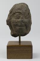 A smiling stone head made of dark terracotta. There are detailed decorative incisions and evidence of hair.
