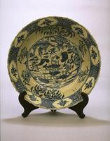 A porcelain large bowl with gently curved interior and straight flaring rim, on a foot ring with kiln grit residue.  It is painted with underglaze cobalt blue to depict a duck in a landscape in the central image surrounded by a border of six groupings of plants, and around the rim eight foliate-shaped reserves frame a floral spray against a patterned ground.  It is covered in a clear glaze with fine crackle.