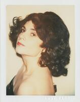 A bust-length portrait of a woman. She is wearing a black dress and red lipstick, and has curly dark hair. She stands with her body turned to the side while she looks into the camera’s lens.
