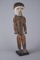 Wooden standing figure with one foot missing. There are two strings of beads around the neck and the face is painted white. The eyes, nose, and mouth are detailed with black pigment. The hair is black with an incised grid-like pattern. 