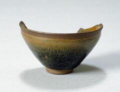 A fragment of a Jian Ware bowl&nbsp;with mottled black-brown glaze. It is on a footing and is dark on the bottom and fades into a lighter color towards the rim.