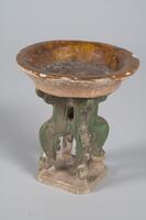 An earthenware miniature amber-glazed basin on a four legged green-glazed stand that mimics wooden furniture, on an unglazed slab base. 