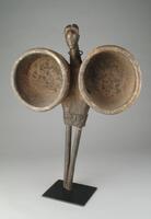 This set of bellows features an elegantly carved male head as a finial. Sitting atop a cylindrical neck that has been pierced with a metal ring, the polished male head features an ovoid face; a high forehead with a vertical line; scarified lines across the cheekbones; and, a coiffure with a finely, detailed pattern at the front and parted down the center.<br />
The man’s “torso” is a trapezoidal piece of wood while two long iron rods functioning as handles represent his “legs.” A pair of round chambers—one on each side of his torso—would originally have had leather bags attached to them, allowing for the pumping of air to heat a fire or forge.