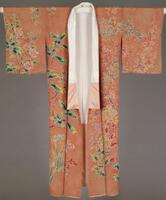 This is a pink silk crepe kimono with wax-resist patterns, hand-painted design and metallic threads embroidery. The kimono is in full length and has elongated sleeves. The fabric is dyed with pink, leaving the family crest under the collar and the floral design part white. The red scale pattern is added using wax-resist technique. Then the design of multiple kinds of plants is hand-painted with white, red, yellow, and pale and blue green colors. There are mix of fall and winter flowers and trees: nandin on the left sleeve, plum, chrysanthemums, thistles, amaranths, camellias and narcissus on the front and back, makino (Chloranthus glaber, with red berries) and more camellias on the right sleeve. Embroidery is added in various metallic threads around the contours of flowers and leaves.