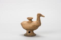 A stoneware vessel designed for pouring or possibly to serve as an oil lamp, in the shape of a duck. The lower half of the duck&#39;s body and &quot;legs&quot; are formed by a shallow bowl on an openwork pedestal; the sides of the bowl have been compressed to make an elongated shape. The upper half of the duck&#39;s body, and its neck and head are formed by hand, The duck&#39;s body is hollow, with two aperture: liquids can be poured in through a funnel with a cup-shaped mouth on the duck&#39;s back, and liquids can be poured out through a wide opening at the tail.<br />
<br />
This is a gray or gray-orange, duck-shaped, low-fired earthenware vessel. Its semi-globular spout is attached to the upper part of the duck&rsquo;s back, while a 2.8cm wide hole, which appears to have been used for pouring liquids, is located at the tail end. The duck&rsquo;s beak is flat and wide, and its eyes are expressed by an incised dot and circle. The lower part of the body features three ridges that form a wave design. The pe