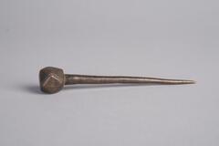 A brass pin with a rounded, diamond shaped head and a long pointed needle on the bottom.