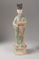 An earthenware sancai three-color glazed figure of a man wearing a long green robe and tall black hat. He is carrying an amber-glazed rectangular box over the top of an amber-glazed tasseled sash that covers his hands, and is standing on a green- and amber-glazed octagonal dais. His face is painted in polychrome mineral pigments, and his head was sculpted separately from the body.