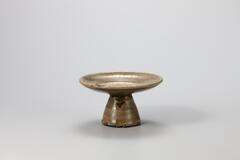 A glazed porcelain offering dish for an altar. The base is a wide cylinder which tapers sharply into a narrow top. The base supports a wide, almost flat disk as the bowl.<br />
<br />
The dish is made from iron-rich clay that tinged the bowl with green-brown. The foot retains traces of sand spurs in eight places. Glaze on the reverse side of the vessel has crawled in parts, revealing the clay body and producing many flaws. This vessel was produced at a regional kiln.<br />
[Korean Collection, University of Michigan Museum of Art (2014) p.200]