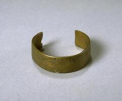 A brass bracelet with a design of central concentric circles edged by triangles. On each side are incised horizontal grooves. Much of the design on the bracelet has worn away. 