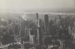 A black-and-white photograph of a cityscape with large skyscrapers, some with plumes of smoke coming from the top, and a river cutting through the distance.