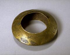 A brass bracelet in the shape of a flattened double cone. There is no decoration on the bracelet, but much of the surface is scratched. 
