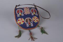 Small, semi-circular beaded bag with three beaded tassels and a braided leather handle. The base of the beadwork is blue, with three faces, two yellow and red flowers, and an interlace pattern decorating the surface. The outside of the bag is edged with yellow and red beads. 