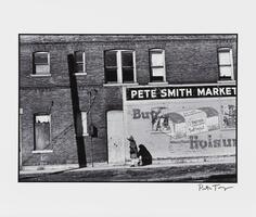 A black and white photograph of a figure wearing a coat and head scarf, walking next to a large brick building. A large sign reads &quot;Pete Smith Market&quot; under which is a bread advertisement.