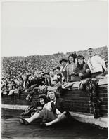 Image of a crowd of people observing something taking place out of frame. A brick wall separates the crowd from two women seated in the foreground.