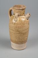 A tall ovoid stoneware ewer with a wide foot and straight neck, an articulated rim, a short straight ribbed spout placed high upon the shoulder, and a coiled handle extending from the mouth to the shoulder.  It is covered in a straw-colored glaze stopping high above the foot.
