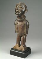 This anthropomorphic <em>nkisi</em>, or power figure, stands upright and features a rather large, forward-tilting head with a prominent, parted mouth, high cheekbones with shallow cheeks, and chipped glass-encrusted lower eyes. A brass tack pierces the figure’s forehead, directly above its nose.  The figure possesses a rectangular torso and robust appendages, although the lower arms and feet are missing. The figure likely held a medicine pack upon its abdomen, evidenced by the four holes bored into its torso, and another one upon its cranium. Around the figure’s neck is a collar, possibly of leather, another place in which medicines are carried.