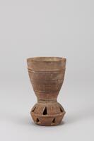 This cup consists of two parts such as the long cup and the midsection containing balls. It has a wide mouth together with a large and flat bottom. Two protruding bands divide the sup into four fields, with the bottom three being decorated with incised gouged dots patterns. The midsection has two areas divided by a protruding band. Both areas have some triangular perforations<br />
<br />
This is a grayish brown, low-fired earthenware bell cup. The bell section is in the shape of two cups attached together at the rim; one cup is upright, while the other cup is reversed. This bell is attached to the base of the cup. The body of the cup is divided into four segments by three sets of raised bands; the lower three segments feature vertical lines of dots. The body of the bell is divided into upper and lower sections, each of which contains a series of triangular perforations offset from those of the other segment. Vertical lines of dots, the same as those on the body, were applied between the perforations in the