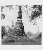 A black-and-white photograph of three large, conical stone structures among trees and remnants of a stone building.