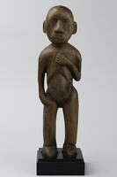 Standing figure with one hand at its hip and the other at its chest. The facial features include an angular nose, slit mouth, and eyes made of glass beads.&nbsp;