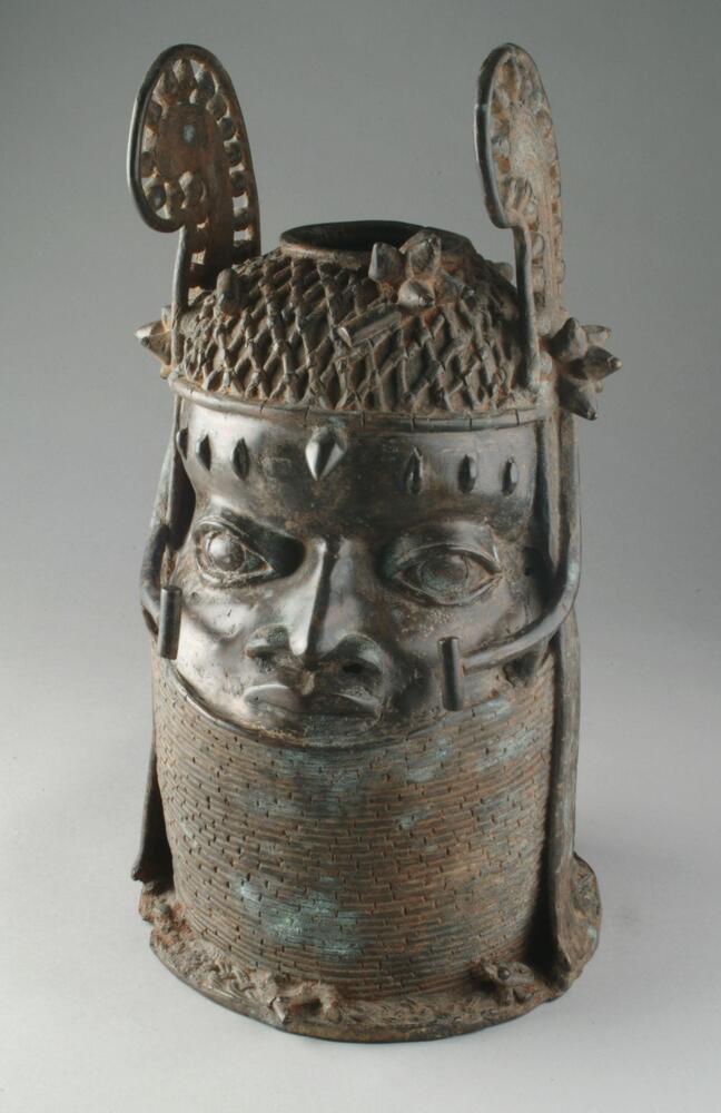 Cast brass head of a human. A beaded collar covers the neck and chin, while the top of the head is decorated with a beaded cap, small clusters of what may be flowers, and 'wings' that rise from either side of the head and almost meet by the mouth. There is an opening at the top of the head as well. 