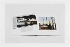 Image of a book lying open to a page featuring a painting of the exterior of smokestacks. A newsclipping featuring a photo of the exterior of smokestacks lies on the book across the open page.  