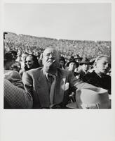 Black and white image of a crowd sitting in stands. A man wearing a suit with a checked shirt and tie, holding a fedora, and smoking a cigar is the in foreground and a boy with blonde hair is on his left.
