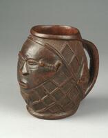 This finely detailed, wooden Wongo cup bears three anthropomorphic faces carved in relief: a large, central face and two smaller, diamond-shaped faces to either side. The remaining areas of the body of the cup are completely covered in an elaborate diamond-shape pattern, a characteristic commonly seen in Wongo and Kuba objects. The cup’s faces exhibit the stylistic influence of the neighboring Kuba, as evidenced by the scarifications on the central face that extend from its temples to its ears; the large, triangular nose; and the half closed coffeebean-shaped eyes. The other two faces closely resemble the center face but lack ears and scarifications. The cup has a curved handle while the interior is smooth and polished.
