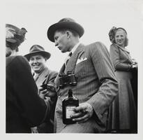 Image of a man in a striped suit and hat. The man holds a bottle with a William Penn label on it and wears binoculars around his neck. Another man and a woman are standing in the background facing the viewer and there is a woman in the left foreground with her back to the viewer.