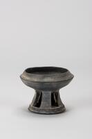 A ceramic rounded bowl with a tall, trapered cylindrical stand with a lip.  The top of the bowl tapers inwards, possibly to allow for a lid to fit it.  The base is detailed with staggered rectangular cut-outs that run from the lip to where the base meets the bowl.