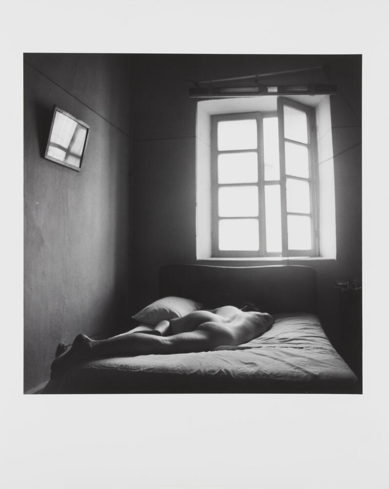 Black and white image of a nude man lying face-down on a bed in front of a window. 