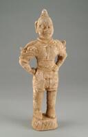 A red earthenware standing figure of a military official or warrior, wearing armor including: a tall pointed helmet, elbow-length gauntlets, a cuirass with plaques, and taces. The armor is worn over a long tunic, loose pants, and boots. The arms are placed upon the hips, and he is standing on a rock-like platform in a contraposto stance, with traces of mineral pigment. 