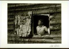 A photograph of a young girl looking out of a window in a log cabin. The inside of the wooden window is pasted with newspaper advertisements.