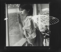 Black and white image of a child looking out a window with white wings superimposed on her back and a white insect watching her.&nbsp;