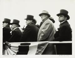 Image of men with their backs turned and looking toward the viewer's left. All are wearing overcoats and hats, with some wearing top hats and flowers in their lapels. 
