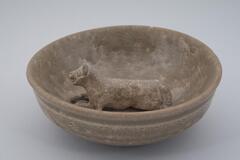 A shallow basin with flat bottom and gently curved sides, a slightly inverted direct rim, and a single reclining dog sculpted in the center of the base.  It is covered in a gray-green celadon glaze.