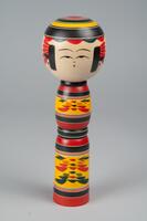 A wooden doll of three tiers made up of a head and body with no arms, legs or feet. Painted on the head is a face, hair, and a yellow, black, and red headdress. The second and third tiers make up the body and have yellow, black, and red decorations.
