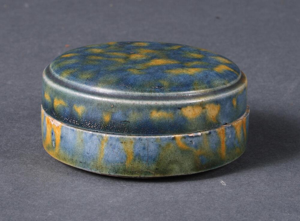 A white earthenware box where the cover and the body are almost identical in form, the entire vessel of a short cylindrical from with flat top, with subtle articulation at the shoulder, on a shallow footring.  It is covered in blue, amber, and green glazes applied in a splotchy manner. 