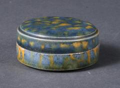 A white earthenware box where the cover and the body are almost identical in form, the entire vessel of a short cylindrical from with flat top, with subtle articulation at the shoulder, on a shallow footring.  It is covered in blue, amber, and green glazes applied in a splotchy manner. 