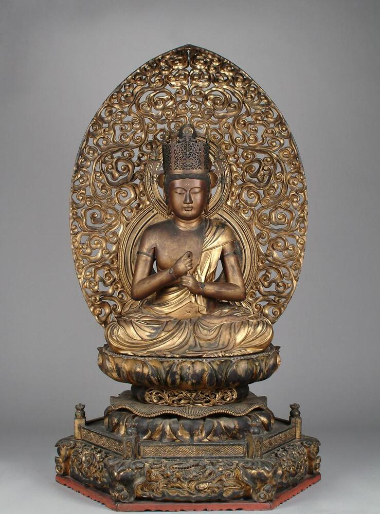A figure is sitting on a lotus-shaped pedestal, which is itself placed on an hexagonal pedestal. The figure wears a drape hanging from the left shoulder and covering the bottom. The arms are placed in front; right hand holding the left index finger. The facial expression is calm; the two eyes looking down; a dot on the forehead. Two elongated ears. A tall crown on the head. The two halos are on the back of the figure; one behind the head and other behind the torso. Two halos are surrounded by an oval-shaped dais. The statue and pedestals are guilded with gold; some polychrome remnants.