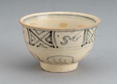 A very small cup of a rounded shape with a slightly everted rim, standing on a relatively high, straight foot. The exterior of the bowl is decorated in two horizontal registers with geometric and abstracted natural motifs drawn with a very free hand in cobalt blue pigment, before a white glaze was applied to the whole cup. The interior of the foot is glazed brown.