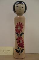 A wooden doll with two tiers made up of a head and body with no arms, legs or feet. Painted on the head is a face and hair. The body is plain except for two flowers painted vertically on the front of the body.